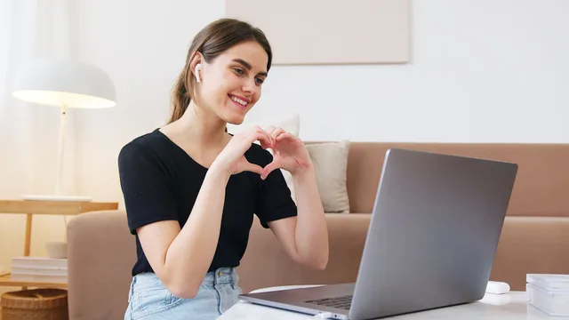 A woman smiling and making a heart with her hands in front of a laptop