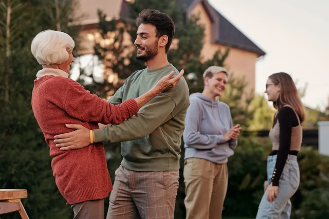 An older woman and a younger man greeting each other with a hug outside a house