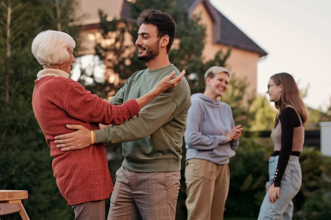 An older woman and a younger man greeting each other with a hug outside a house